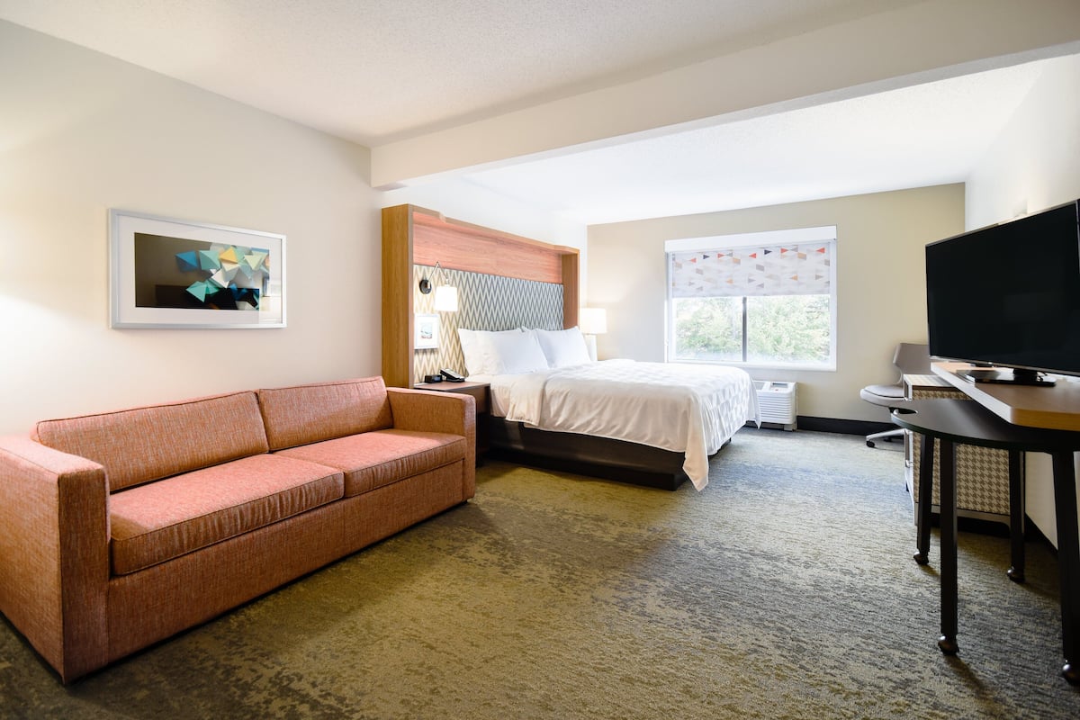 A hotel room with a bed, a sofa, a desk, a television, and a window with blinds.