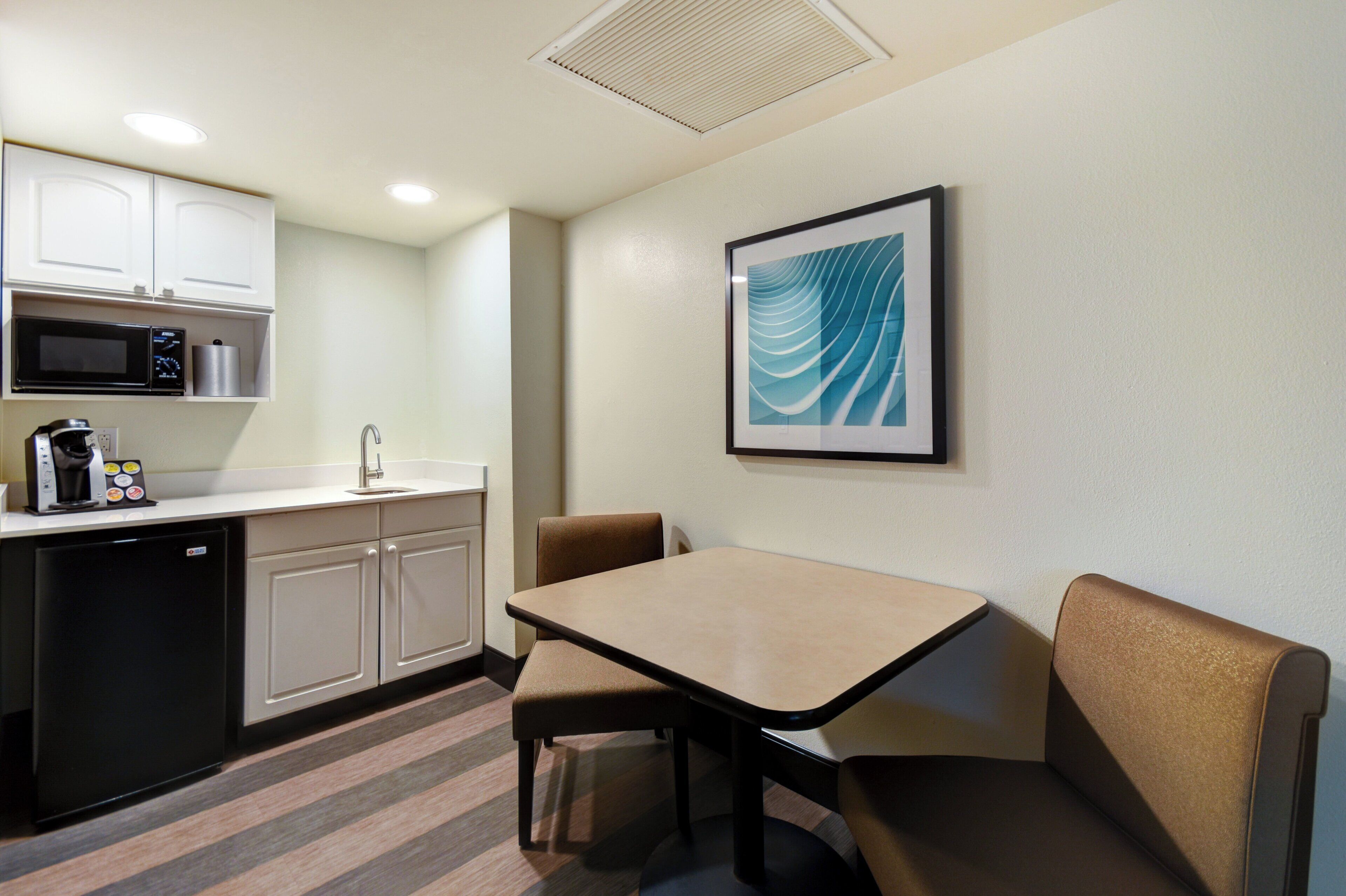 Una habitación de hotel compacta con kitchenette, una mesa de comedor con dos sillas y una obra de arte enmarcada en la pared.