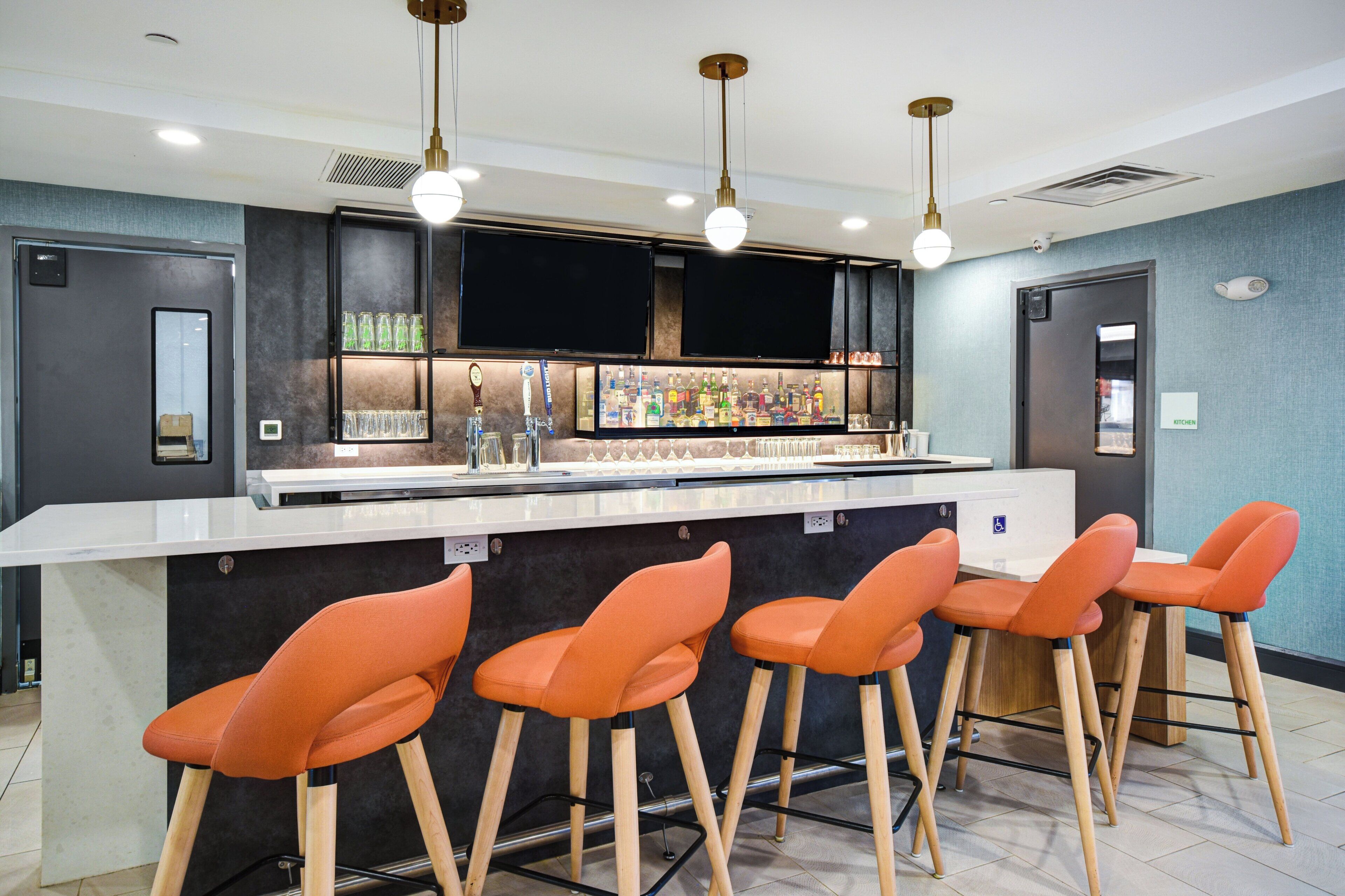 Zona de bar con mostrador blanco, varios taburetes y una variedad de botellas y vasos.