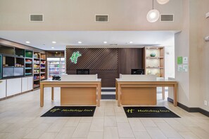 L’espace d’accueil d’un hôtel Holiday Inn, doté d’un design moderne, comprend deux comptoirs d’accueil, une grande enseigne lumineuse et divers équipements, notamment un réfrigérateur rempli de boissons et d’en-cas.