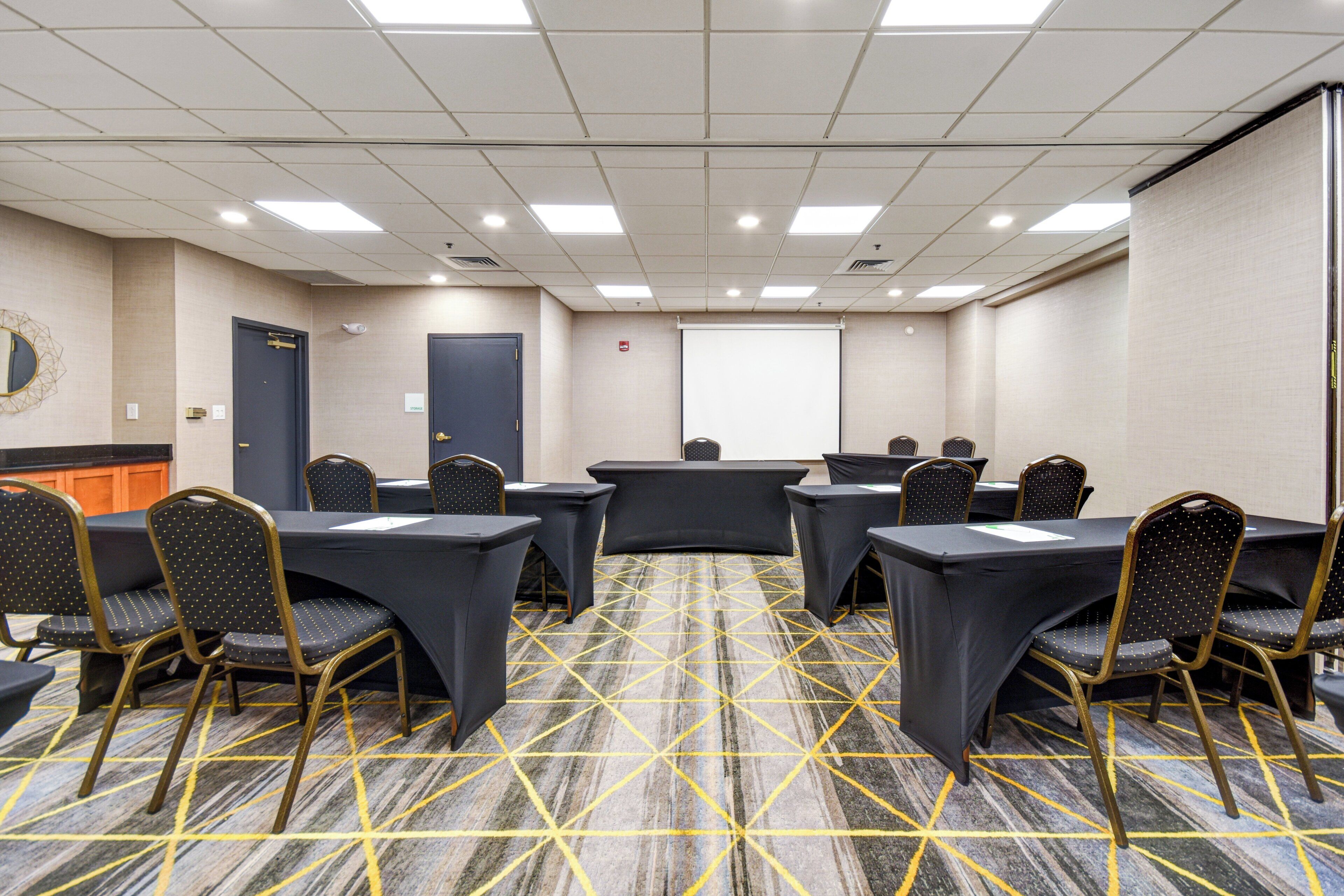 A conference room with a carpeted floor, tables, chairs, a projector screen, and a door.