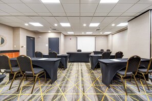 Une salle de conférence dotée d’un sol recouvert de moquette, de tables, de chaises, d’un écran de projection et d’une porte.