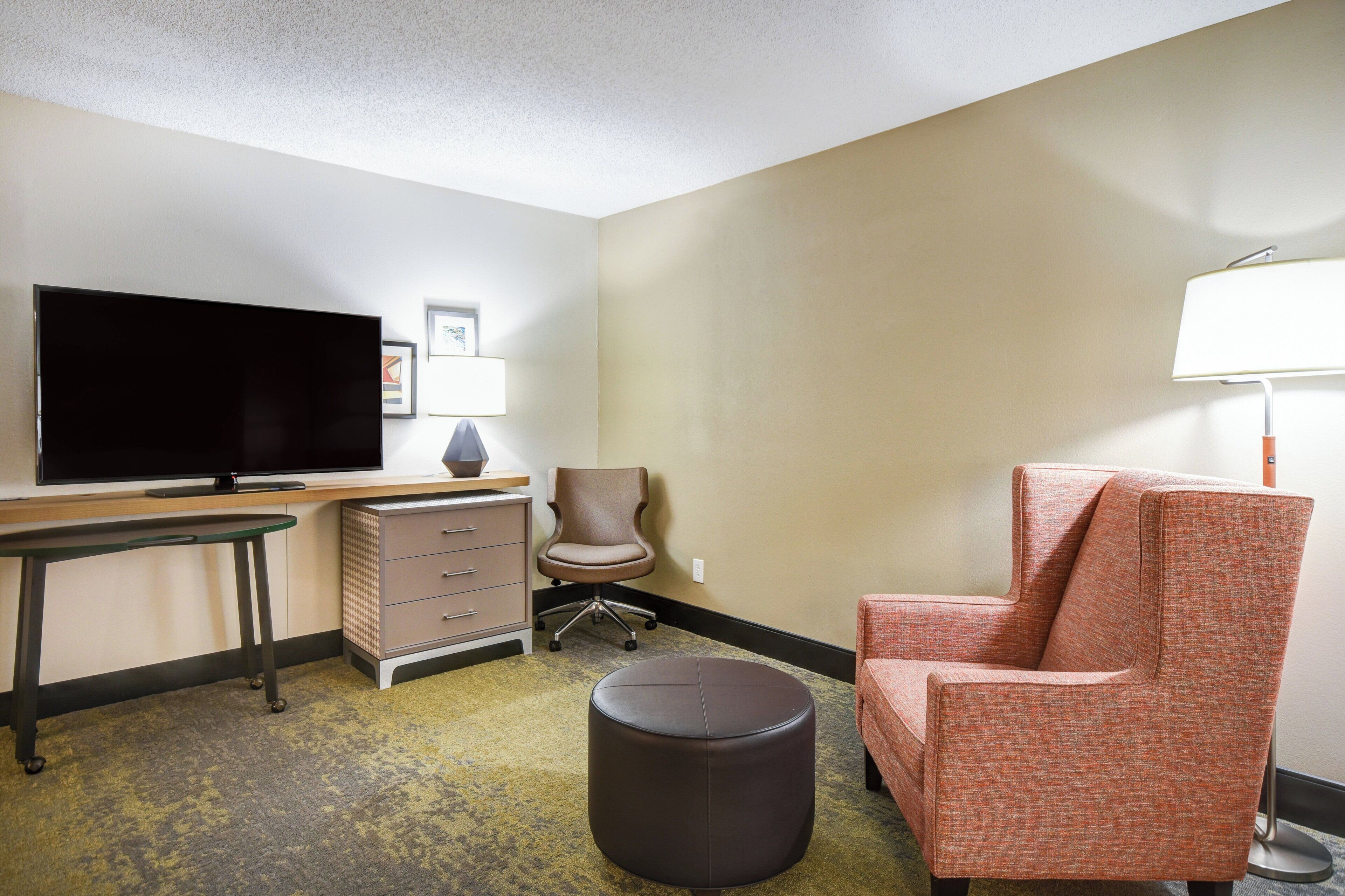 A hotel room with a flat-screen TV, a desk, a chair, an ottoman, and a lamp.
