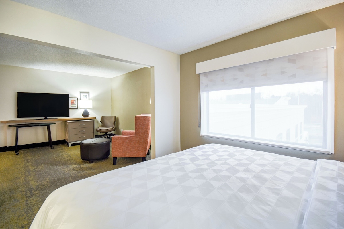 Ein Hotelzimmer mit Bett, Schreibtisch, Stuhl, Fernseher und einem Fenster mit Jalousien.