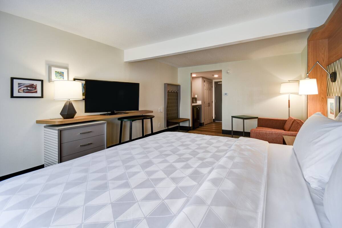 A hotel room with a bed, a television, a desk with a lamp, a chair, and a sofa.