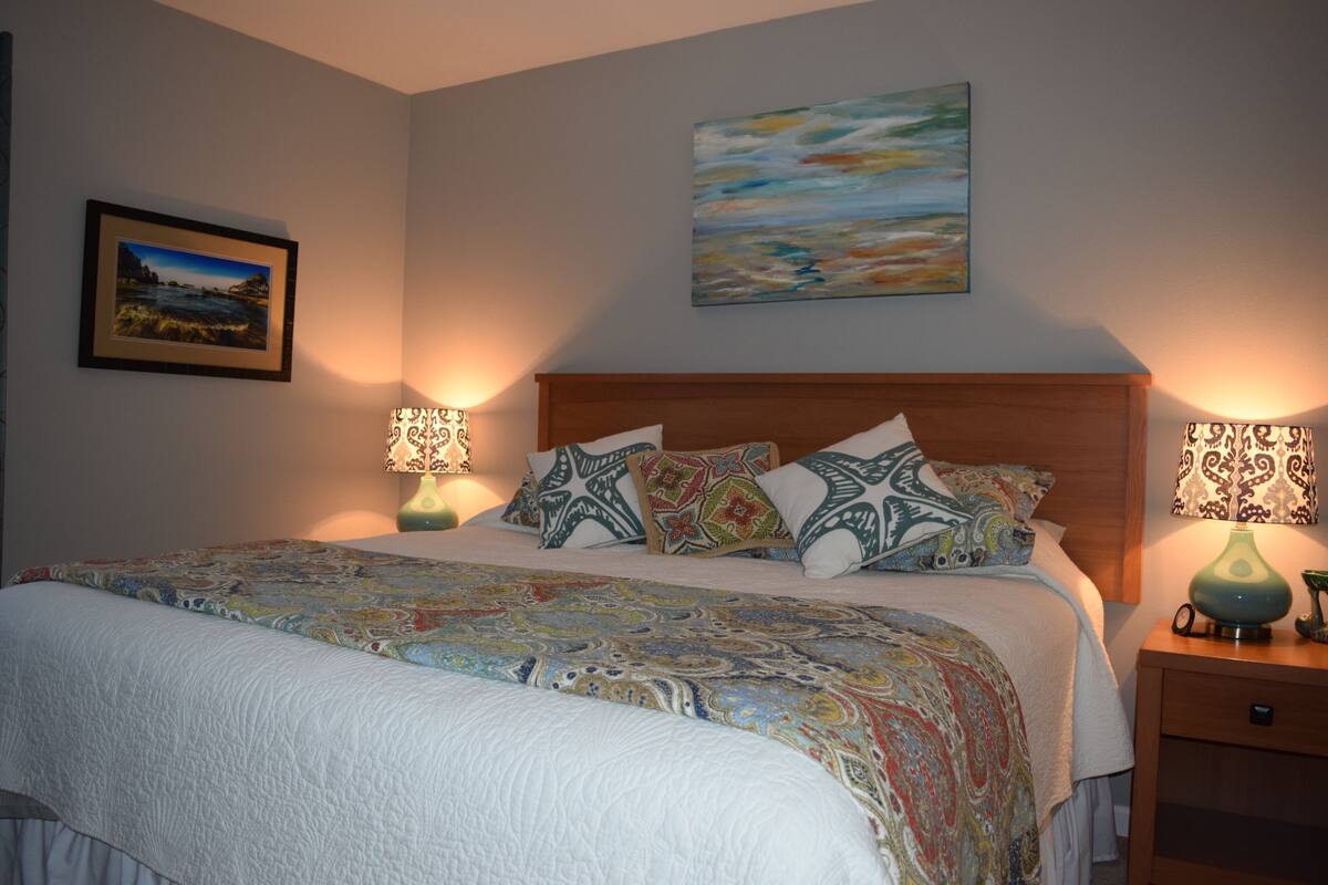 A neatly made bed with patterned bedding, two bedside lamps, and a framed landscape painting on the wall.