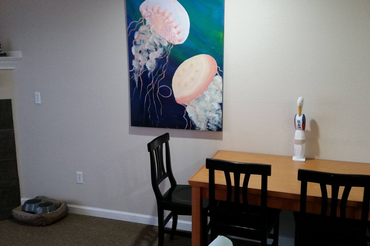 A dining area with a wooden table and four chairs, a painting of jellyfish on the wall, and a small statue of a bird on the table.
