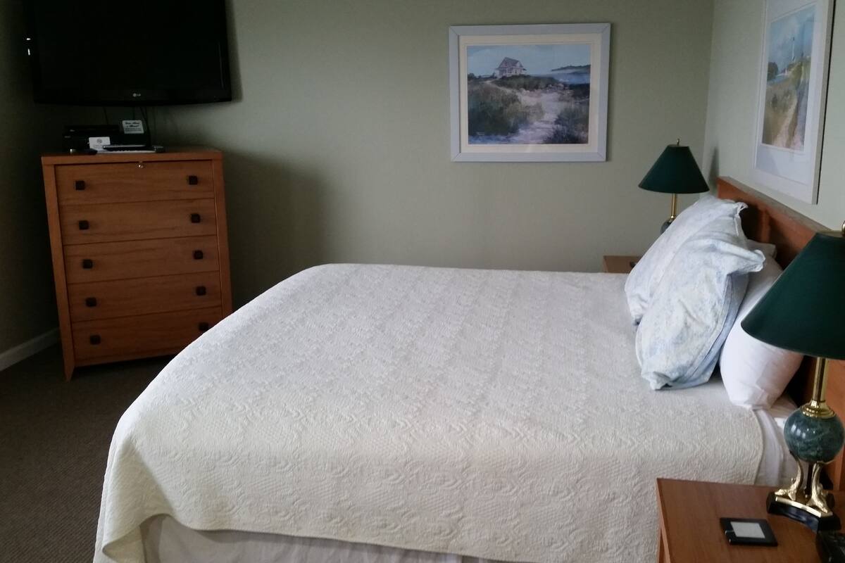 A neatly made bed with a white comforter, a wooden dresser with a flat-screen TV, and two wall-mounted lamps.