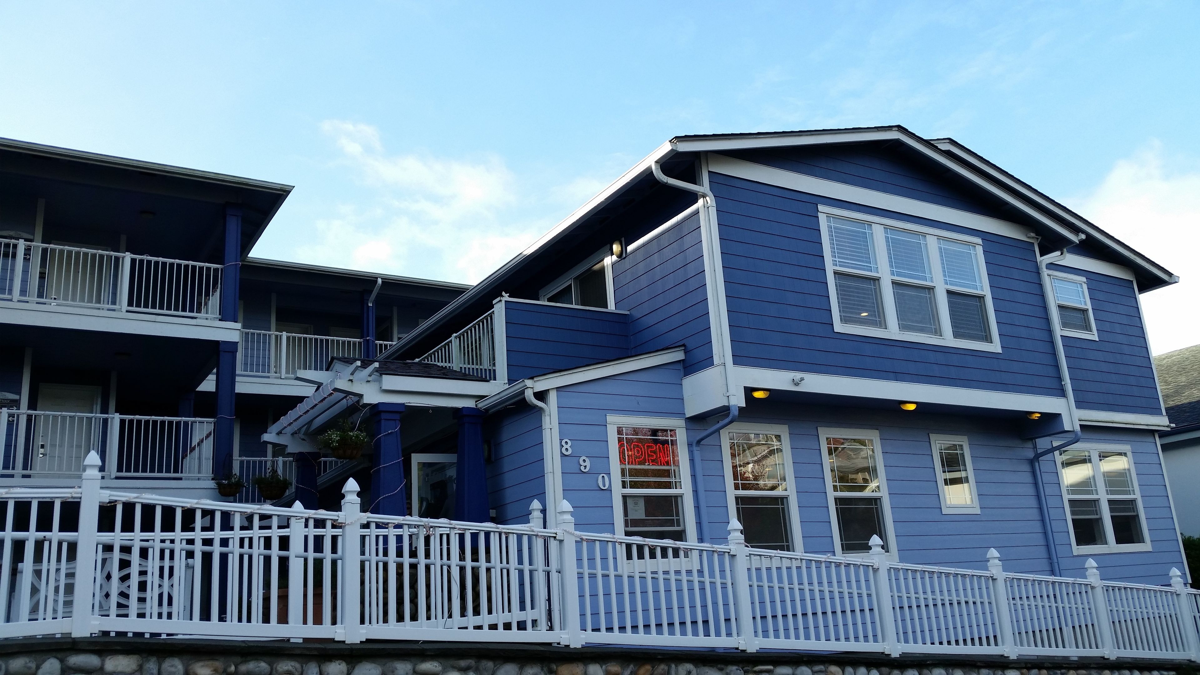 A two-story building with a blue exterior and white trim, featuring a balcony on the upper floor and a white picket fence in the foreground.