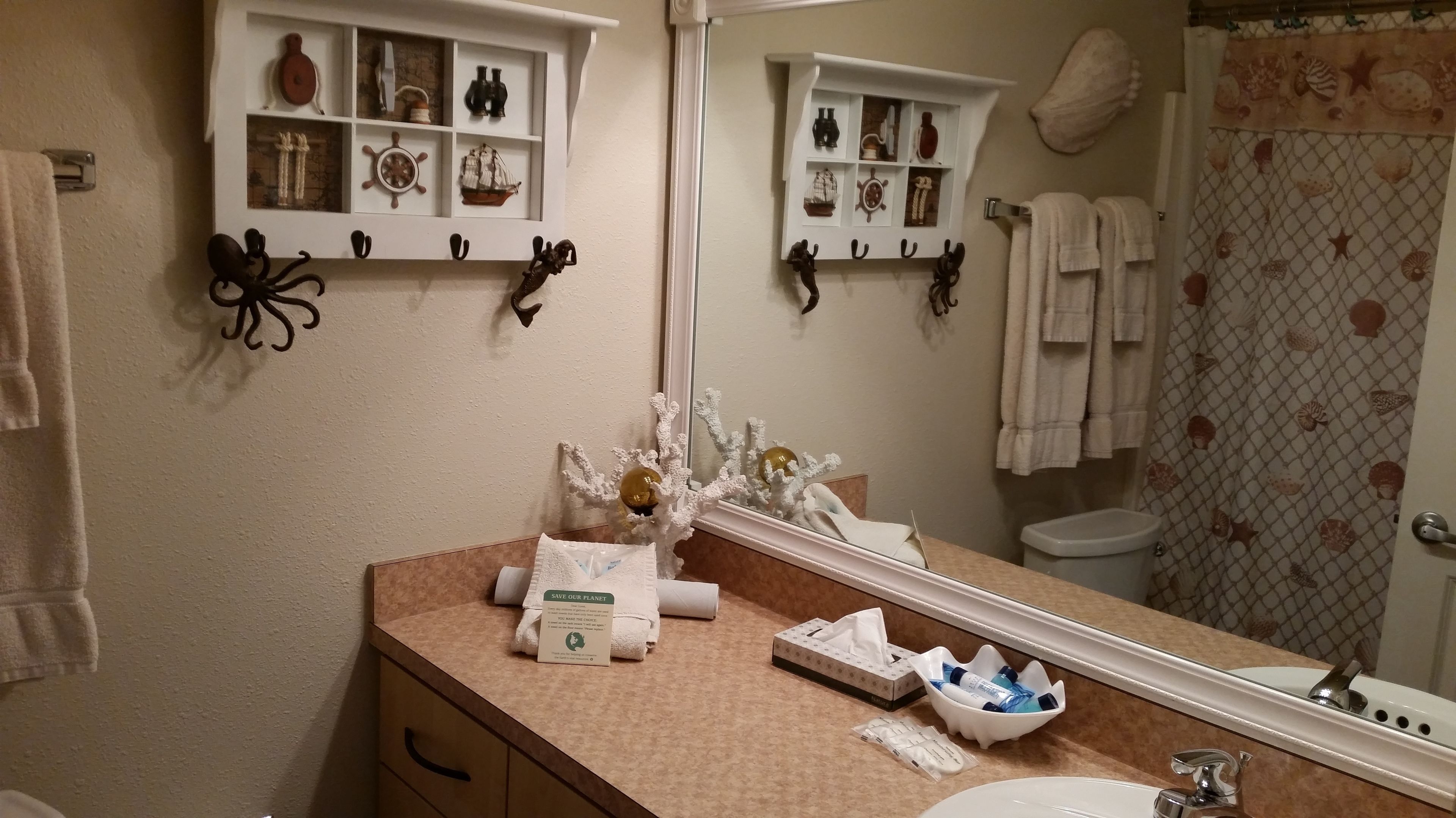 A bathroom with a beige countertop, a sink, a mirror, and shelves with nautical-themed decorations.