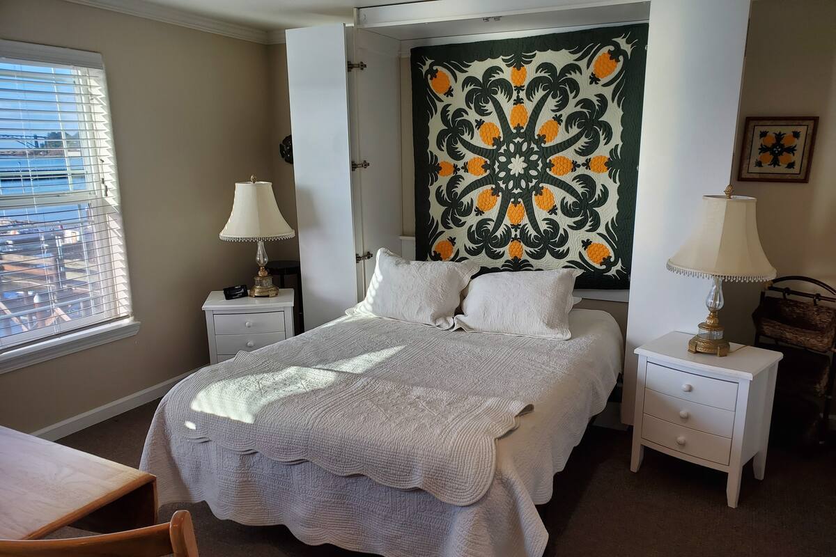 A bedroom with a bed, two nightstands, a window with blinds, a chair, and a view of a bridge.