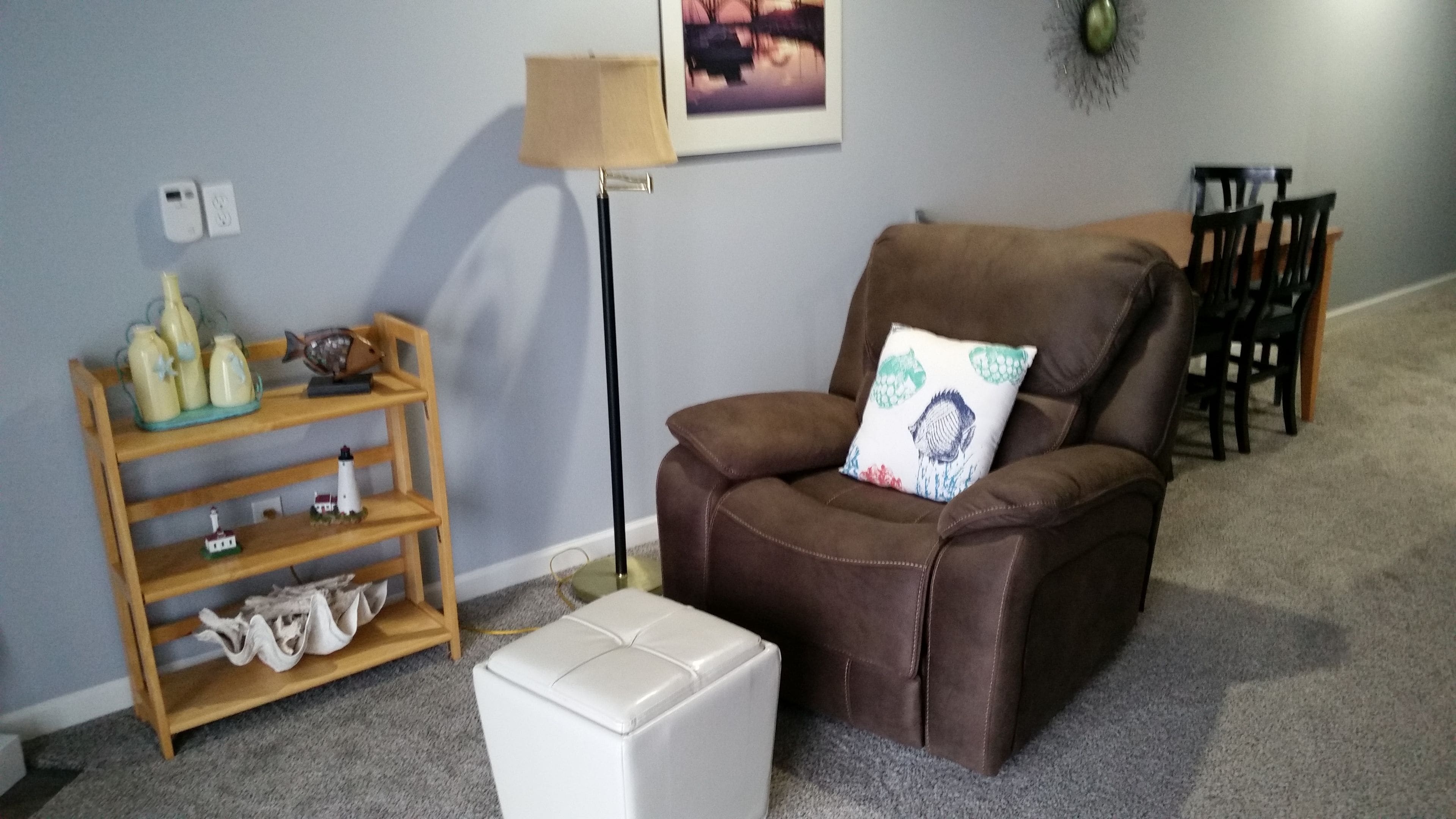 Uma sala de estar com uma poltrona de couro marrom, um banquinho branco, uma estante de madeira com objetos decorativos, um abajur e um quadro na parede.