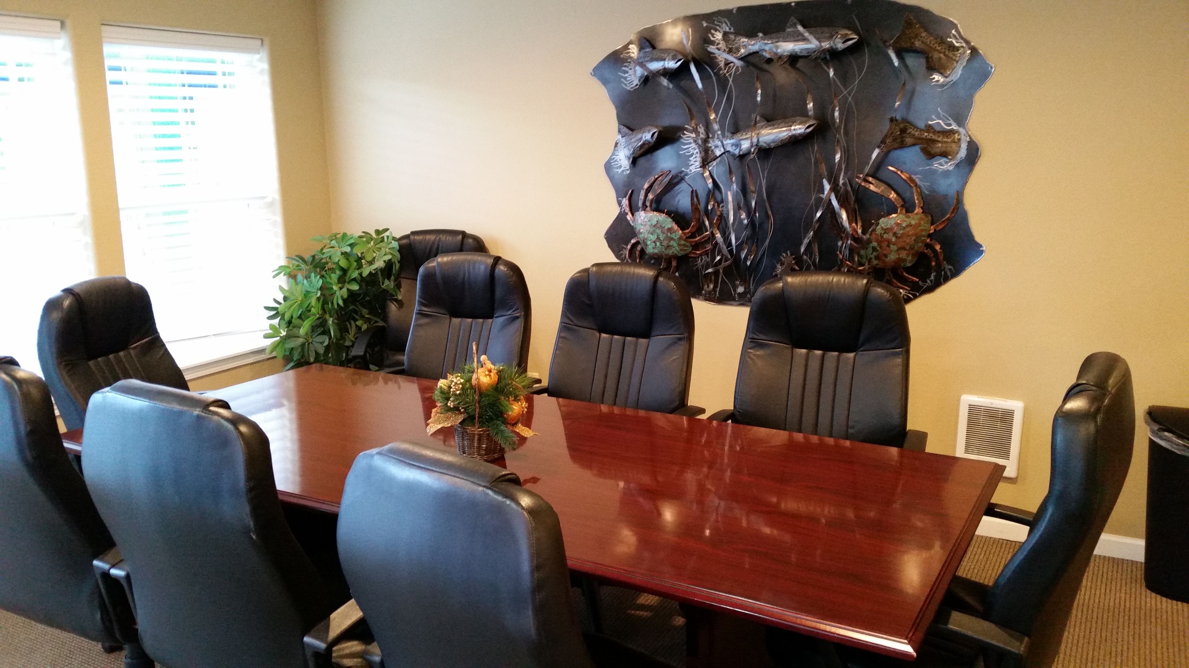A conference room with a wooden table, six black leather chairs, a decorative wall piece, and a potted plant.