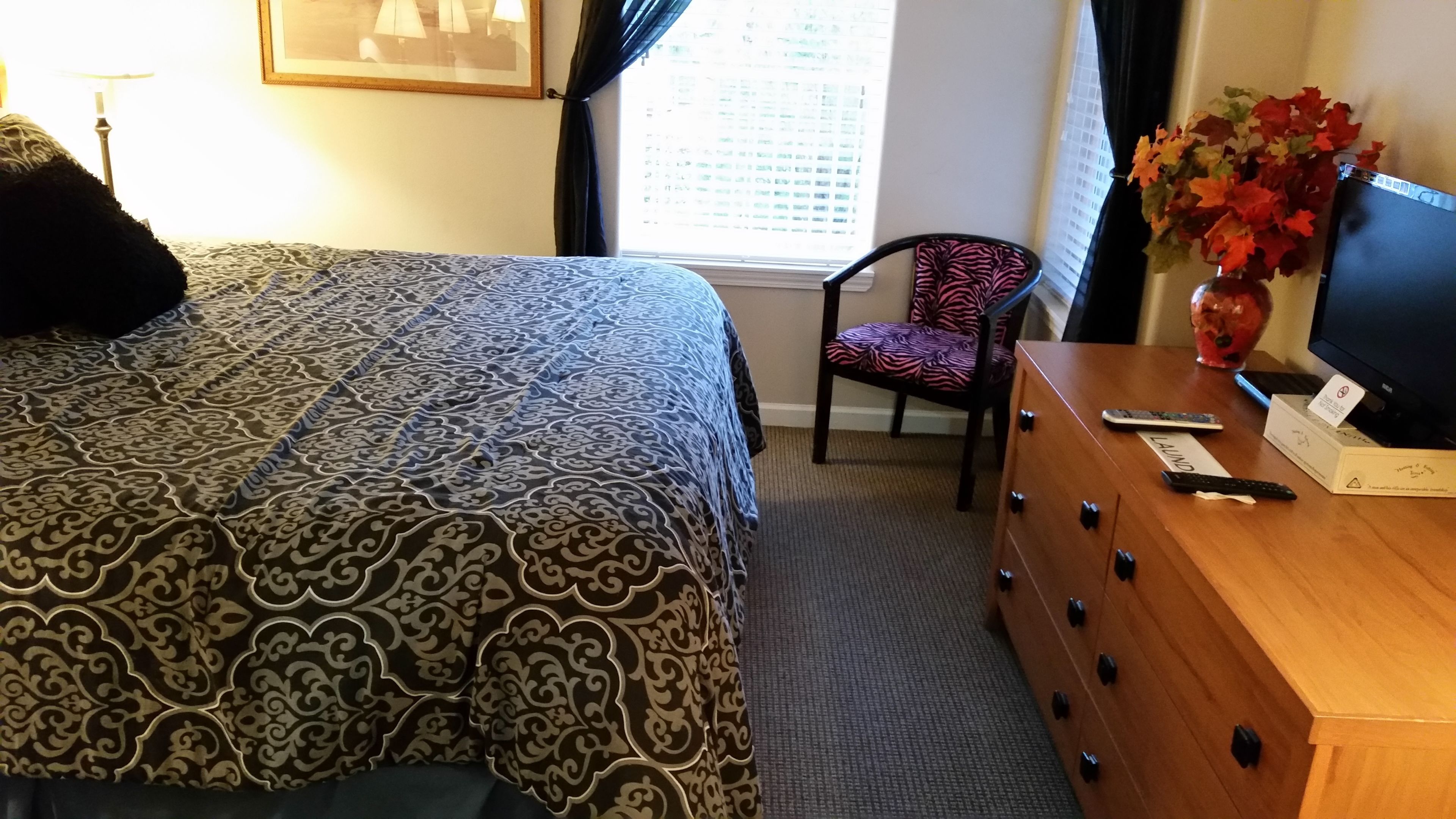 Une chambre d’hôtel avec une couvre-lit à motifs, une commode en bois surmontée d’un téléviseur, une chaise et un vase rempli de fleurs.