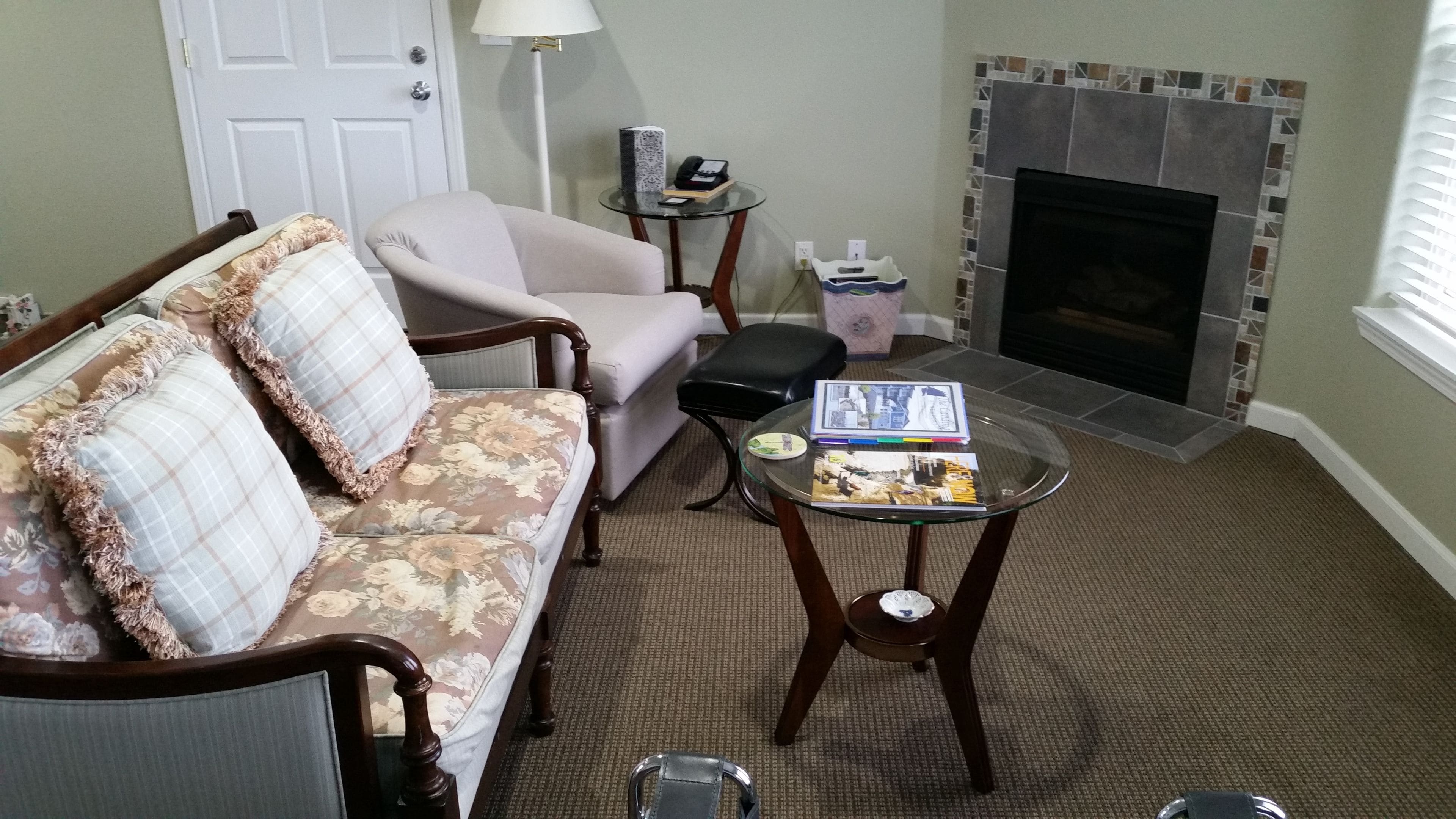 A living room with a sofa, armchair, and a small table with magazines. There is a fireplace and a door in the background.