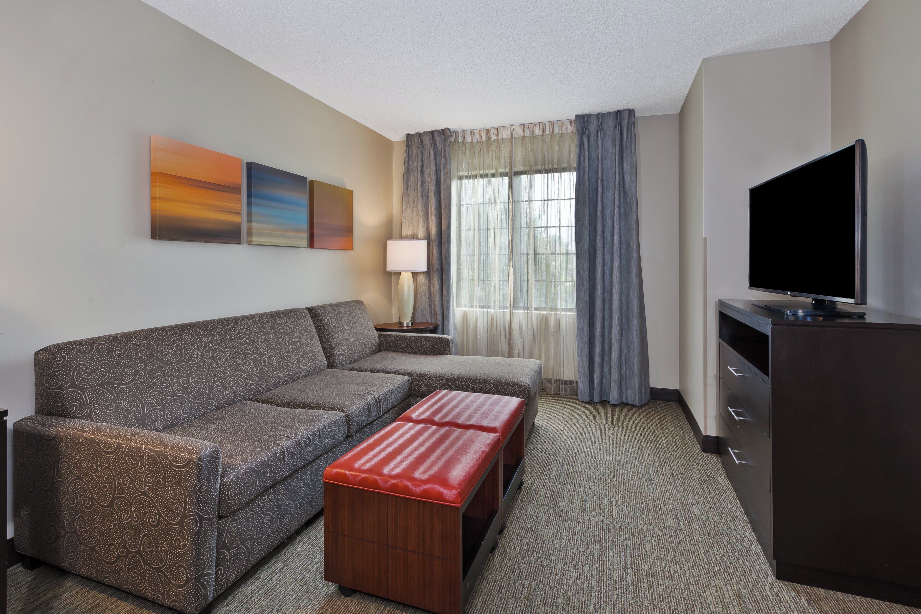 A hotel room with a sofa, a small table, a lamp, a TV, and a window with curtains.
