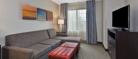A hotel room with a sofa, a small table, a lamp, a TV, and a window with curtains.