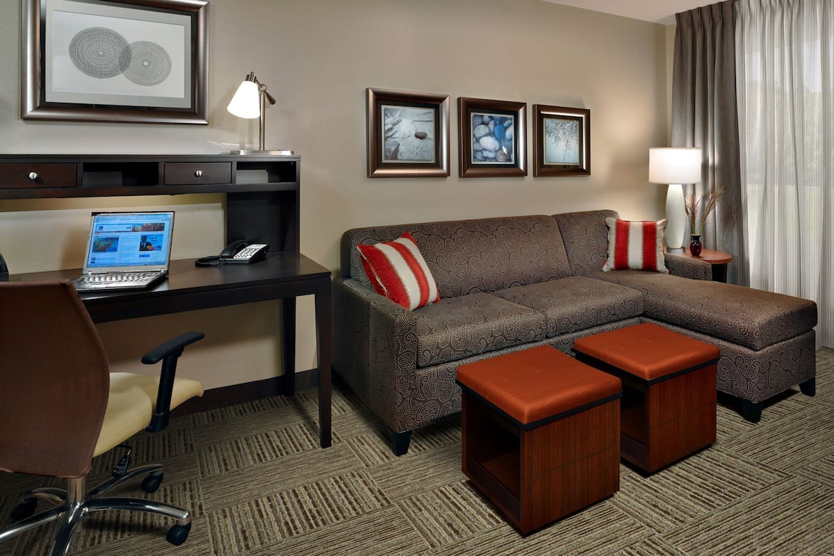 A hotel room with a brown sectional sofa, a desk with a laptop, a telephone, and a TV. There are two ottomans, a lamp, and framed artwork on the wall.