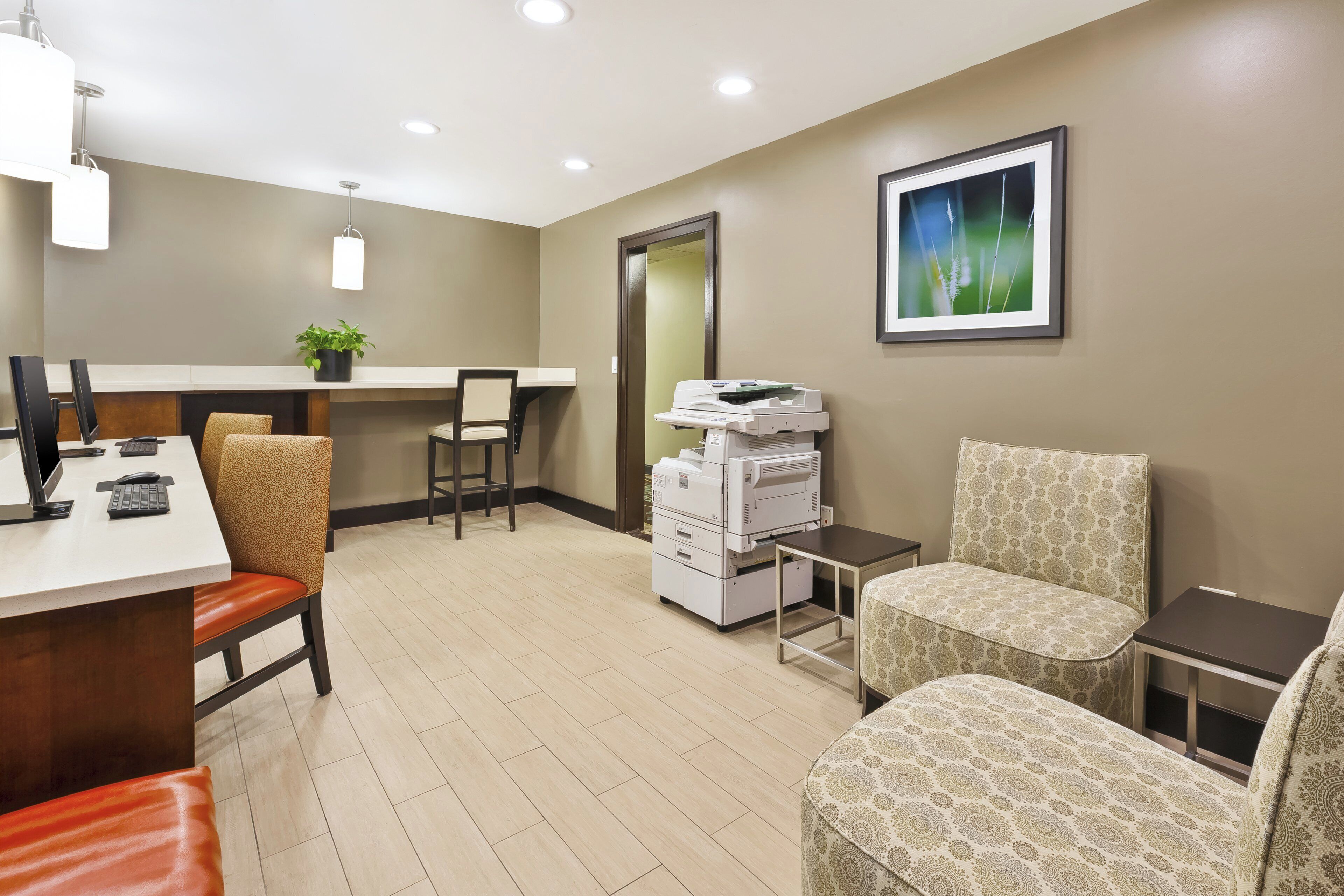 A hotel room with a desk, chairs, a printer, a potted plant, and a framed picture on the wall.