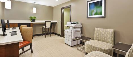 A hotel room with a desk, chairs, a printer, a potted plant, and a framed picture on the wall.