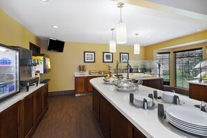 A hotel kitchen with a buffet setup, including a refrigerator stocked with beverages, a flat-screen TV mounted on the wall, and a large window with blinds. - Staybridge Suites Detroit-Utica by IHG (Utica)