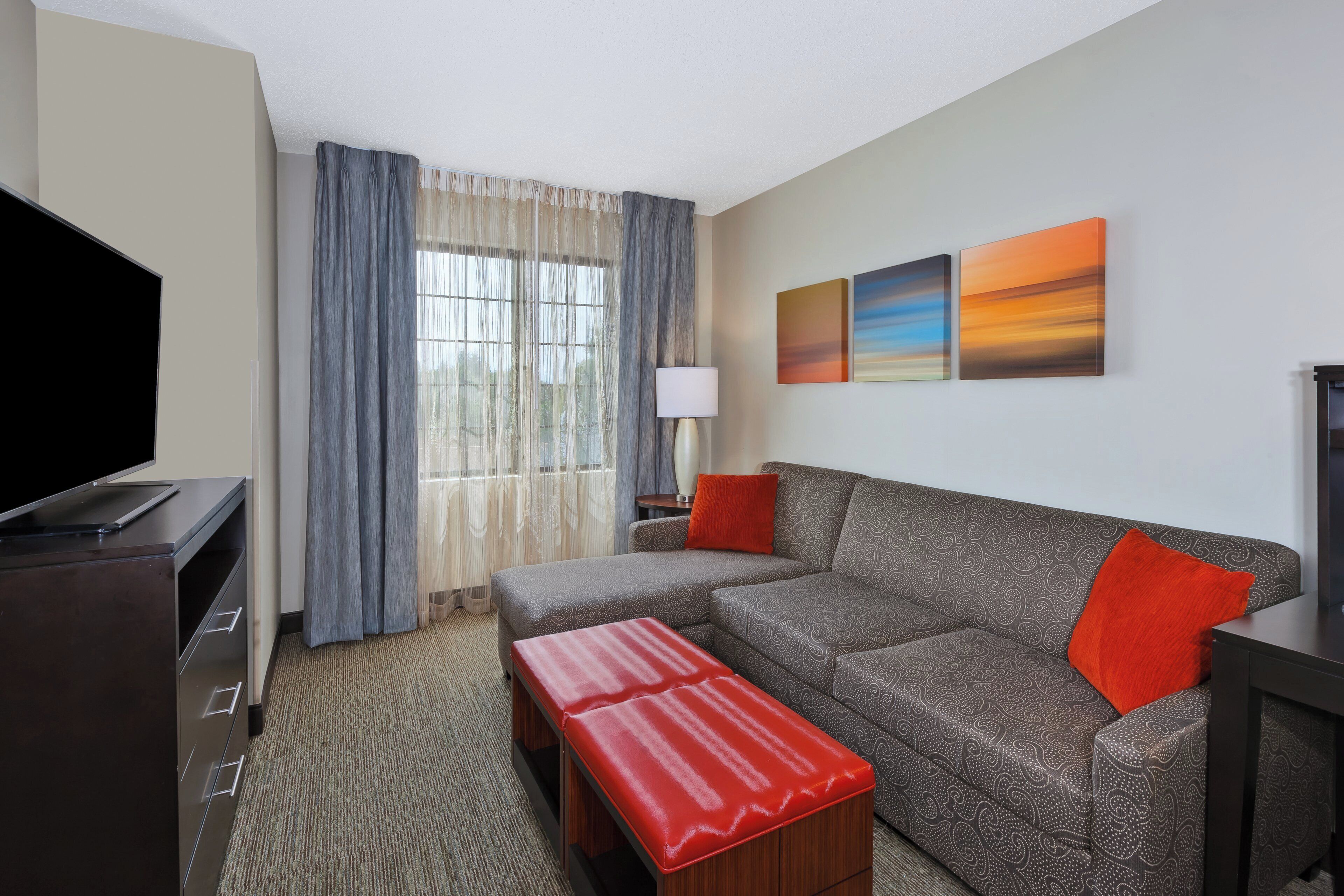 A hotel room with a sofa, a red ottoman, a TV, and artwork on the wall.