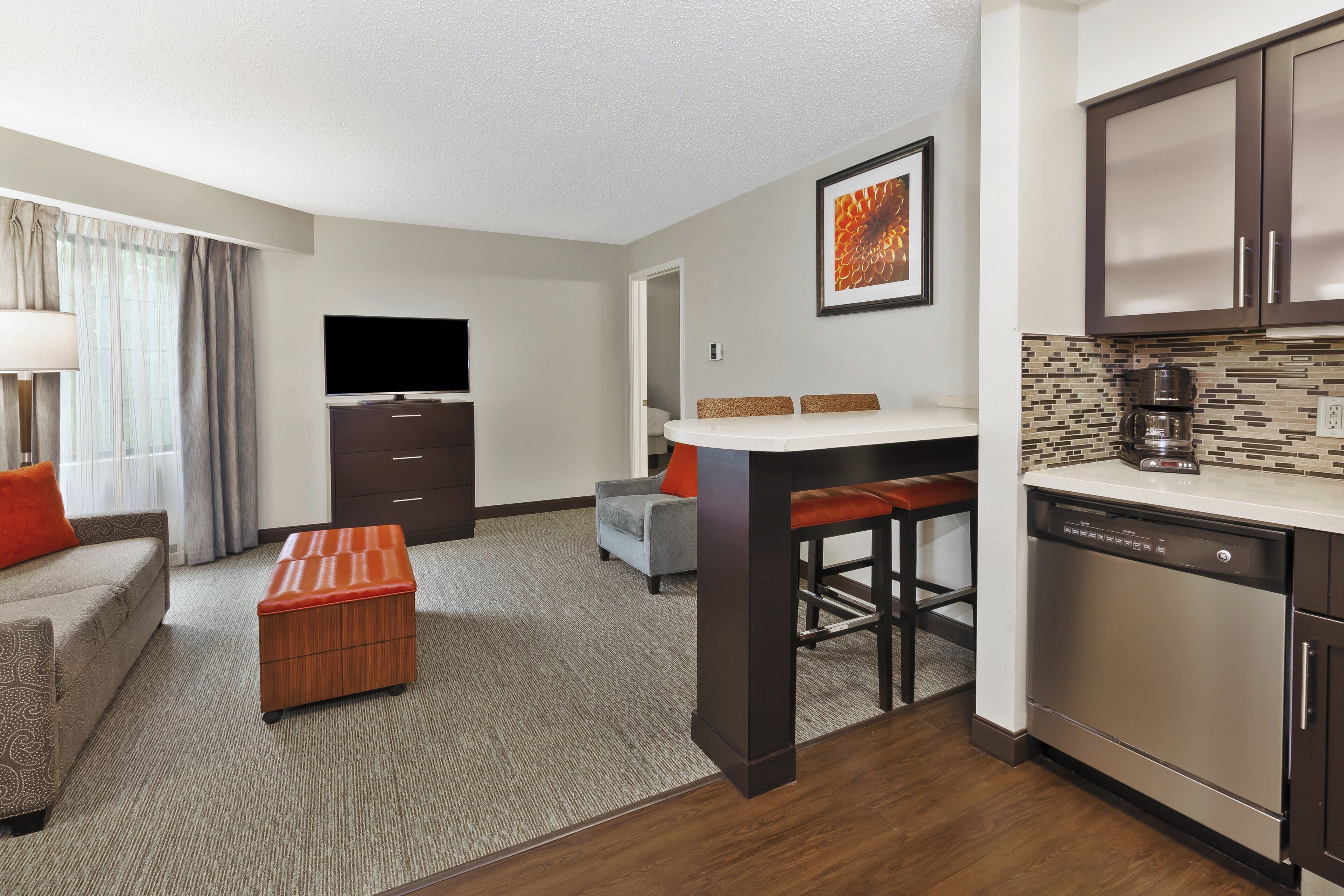 A hotel room with a sofa, ottoman, TV, kitchenette, and a dining area.