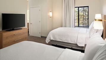 Une chambre d’hôtel avec un téléviseur à écran plat posé sur une commode en bois, deux lits avec du linge de lit blanc, une fenêtre donnant sur des arbres et une lampe fixée au mur.