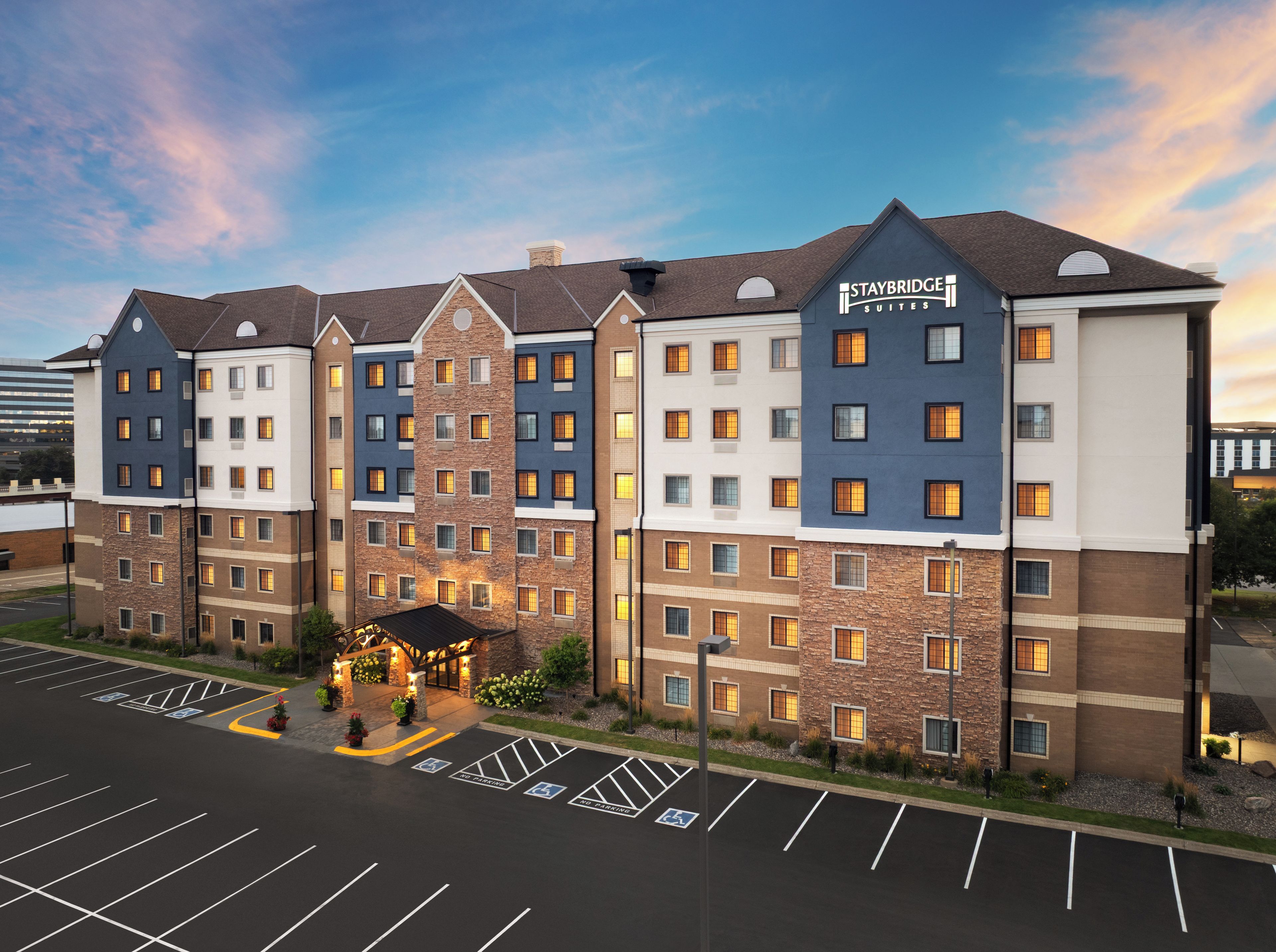 A multi-story hotel with Staybridge Suites branding, featuring a brick facade, multiple balconies, and a parking lot with handicapped accessible spaces.