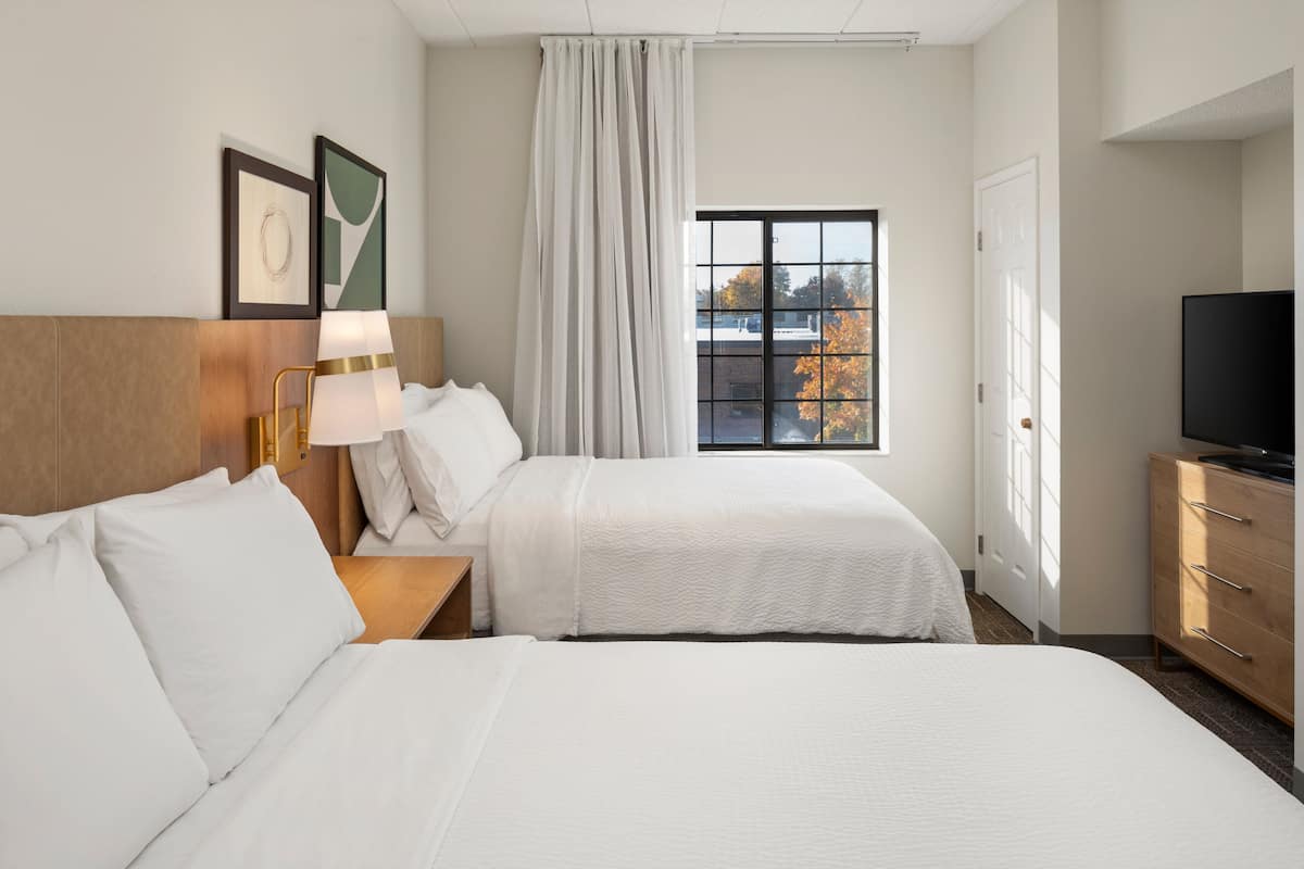 A hotel room with two beds, a TV, a desk, and a window with a view of trees.
