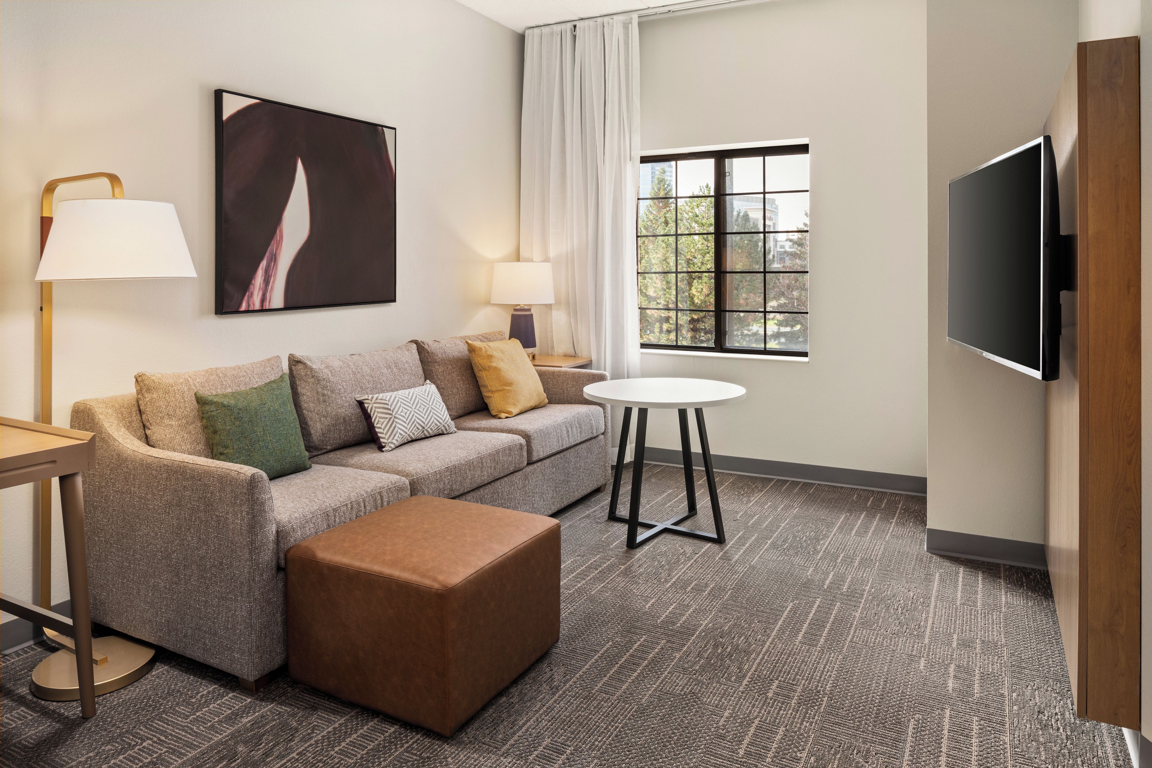 Habitación de hotel con sofá, reposapiés, dos lámparas, televisor y ventana con vista a árboles.