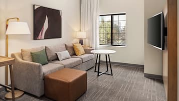 Habitación de hotel con sofá, reposapiés, dos lámparas, televisor y ventana con vista a árboles.