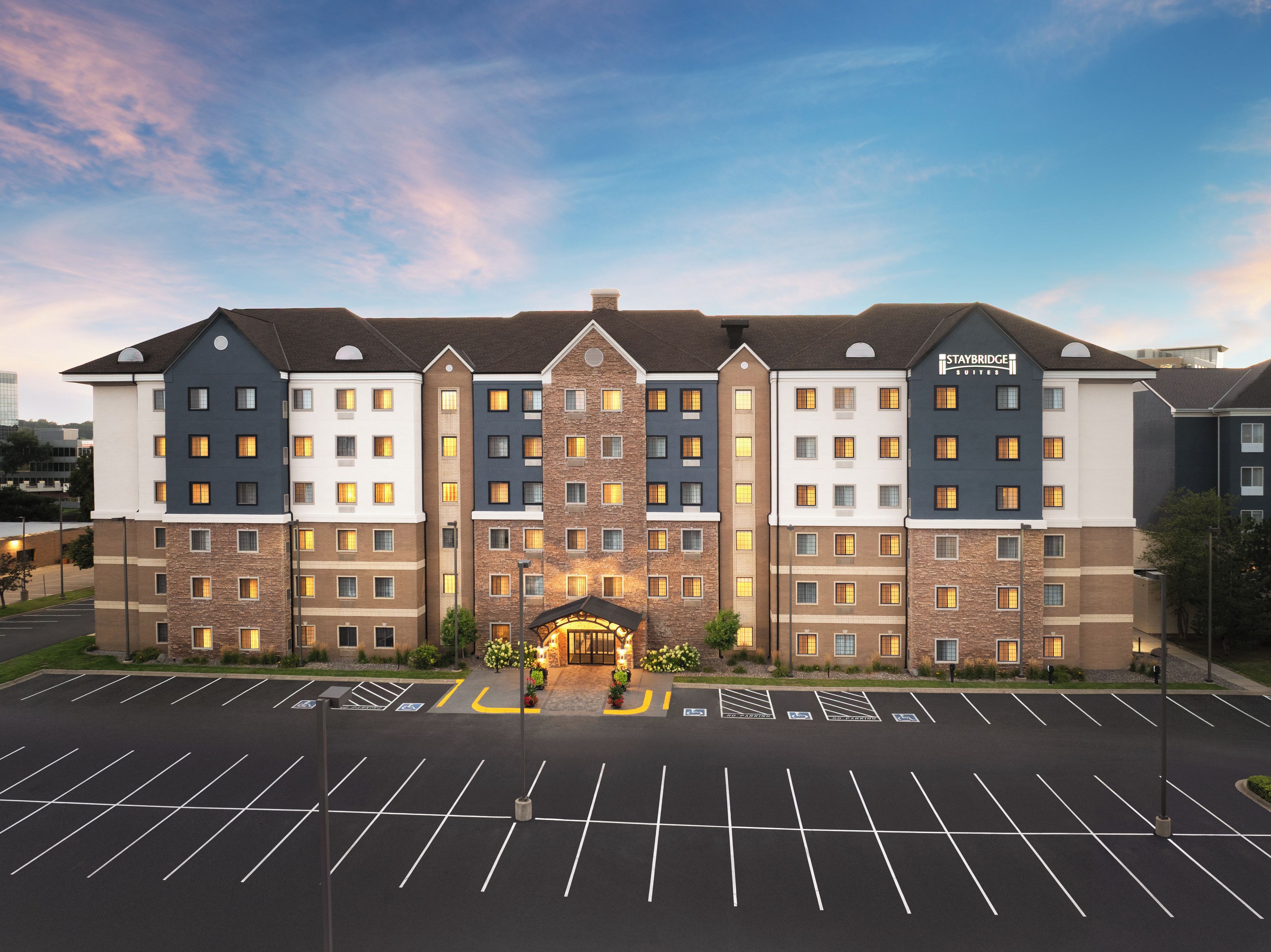 A multi-story hotel with a central entrance, parking lot, and a clear sky.