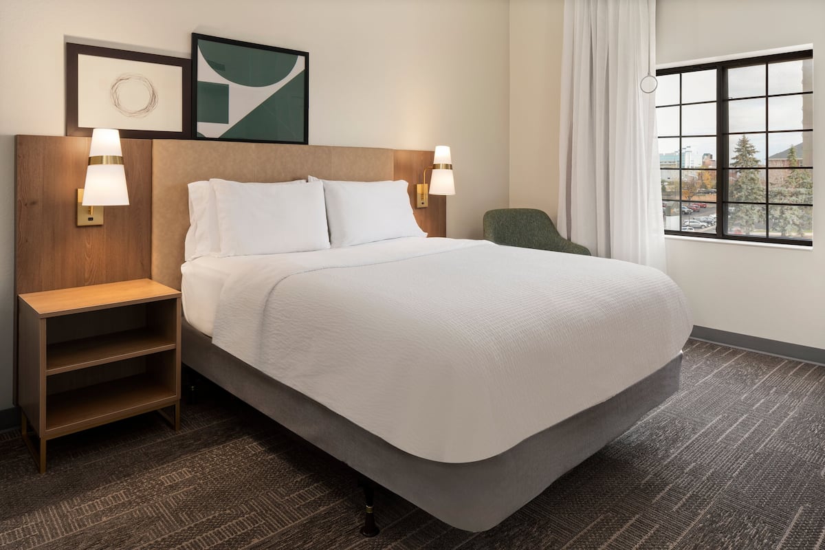 A neatly made bed with a wooden headboard, two bedside lamps, a small wooden nightstand, and a view of the outside through a window.