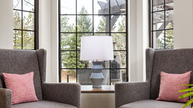 A corner seating area with two grey armchairs and a white lamp, positioned in front of a large window with a view of trees.
