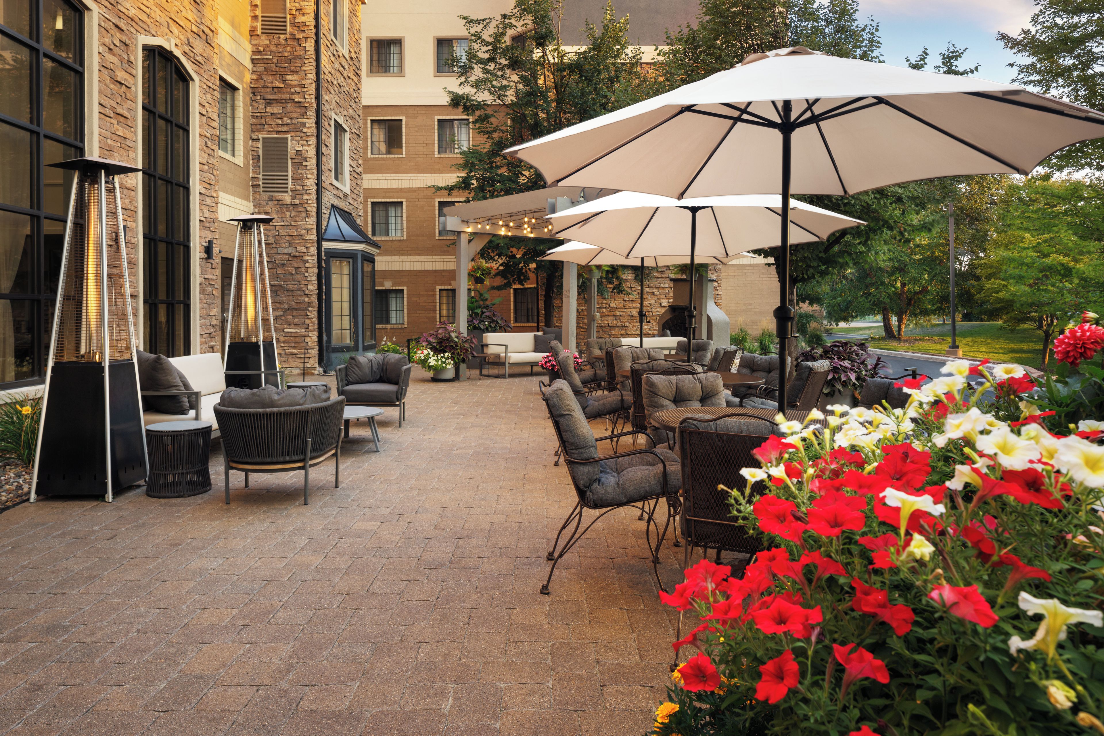 An outdoor patio area with brick flooring, multiple seating arrangements including sofas and chairs, and large umbrellas providing shade.