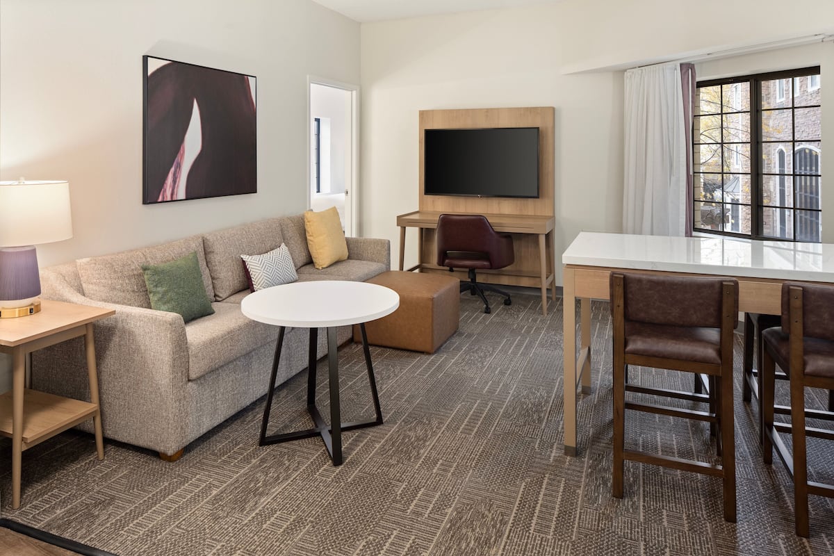 A modern hotel room with a sofa, a small round table, a TV mounted on a wooden panel, and a dining area with chairs.