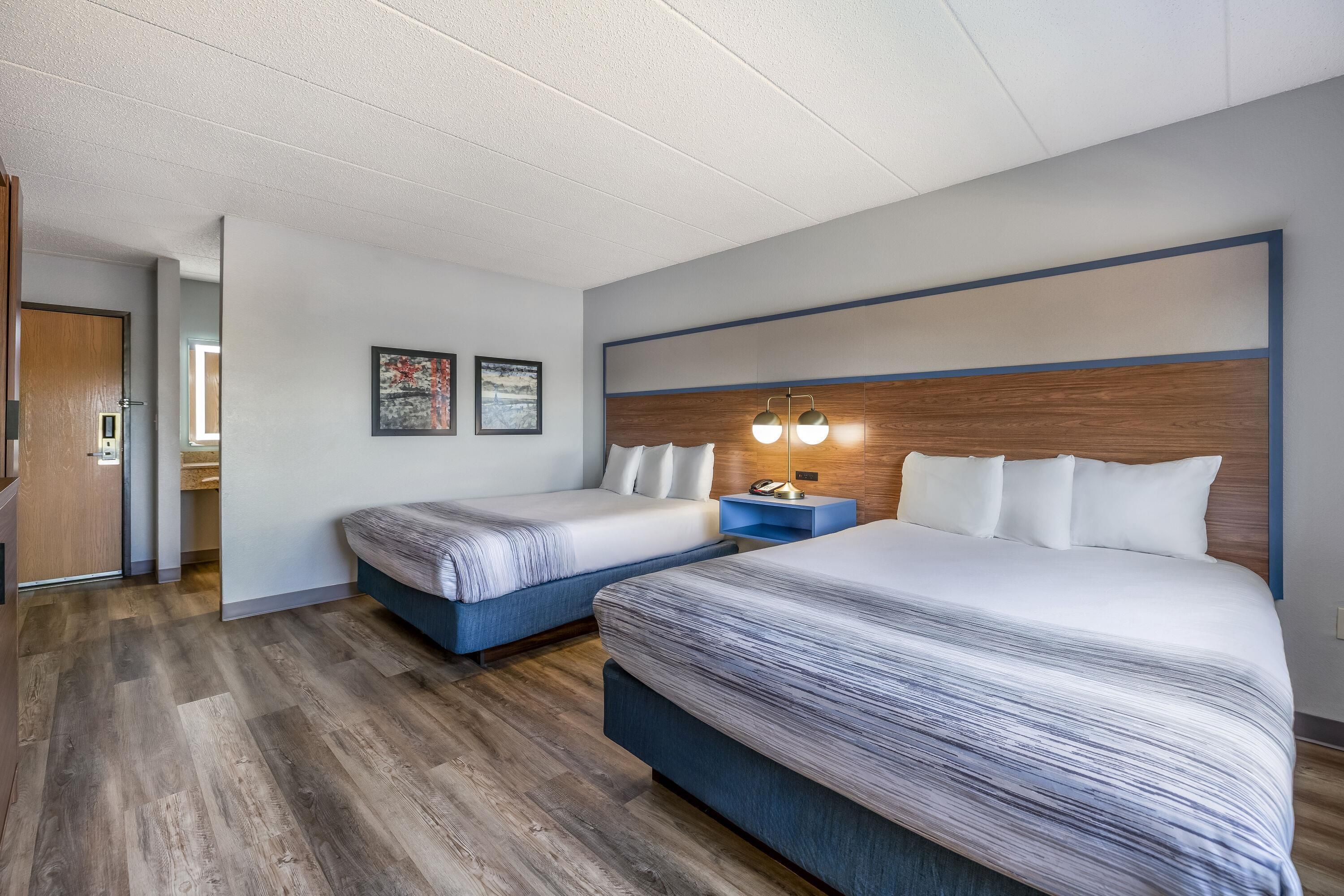 A hotel room with two beds, a wooden headboard, a nightstand with a lamp, and a door leading to a bathroom.