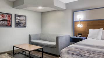A hotel room with a bed, a grey armchair, a small table, a nightstand with a phone, and two wall-mounted artworks.