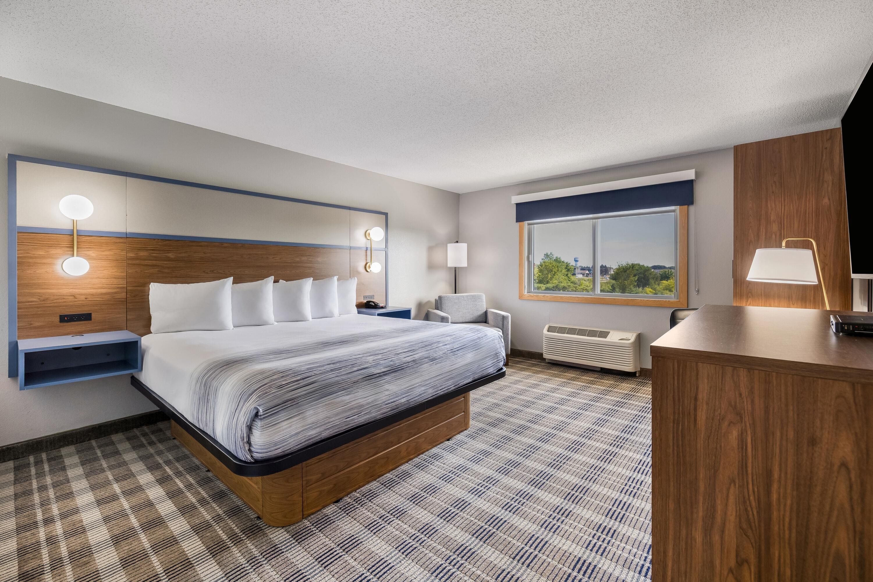 A hotel room with a bed, bedside tables, a desk, a chair, a window with a view, and a built-in heater.