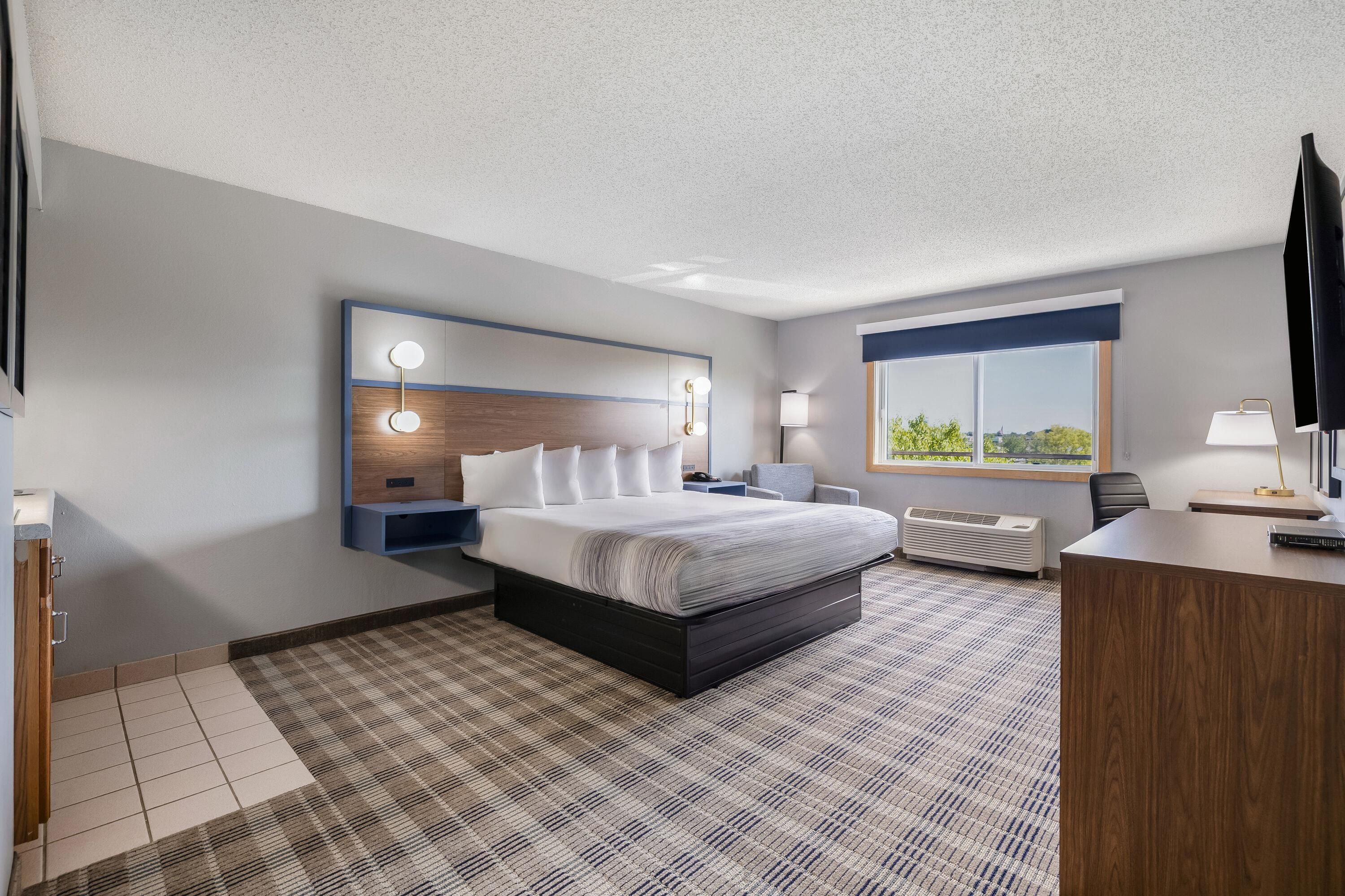 A hotel room with a bed, bedside tables, a desk, a chair, a window with a view, and a flat-screen TV.