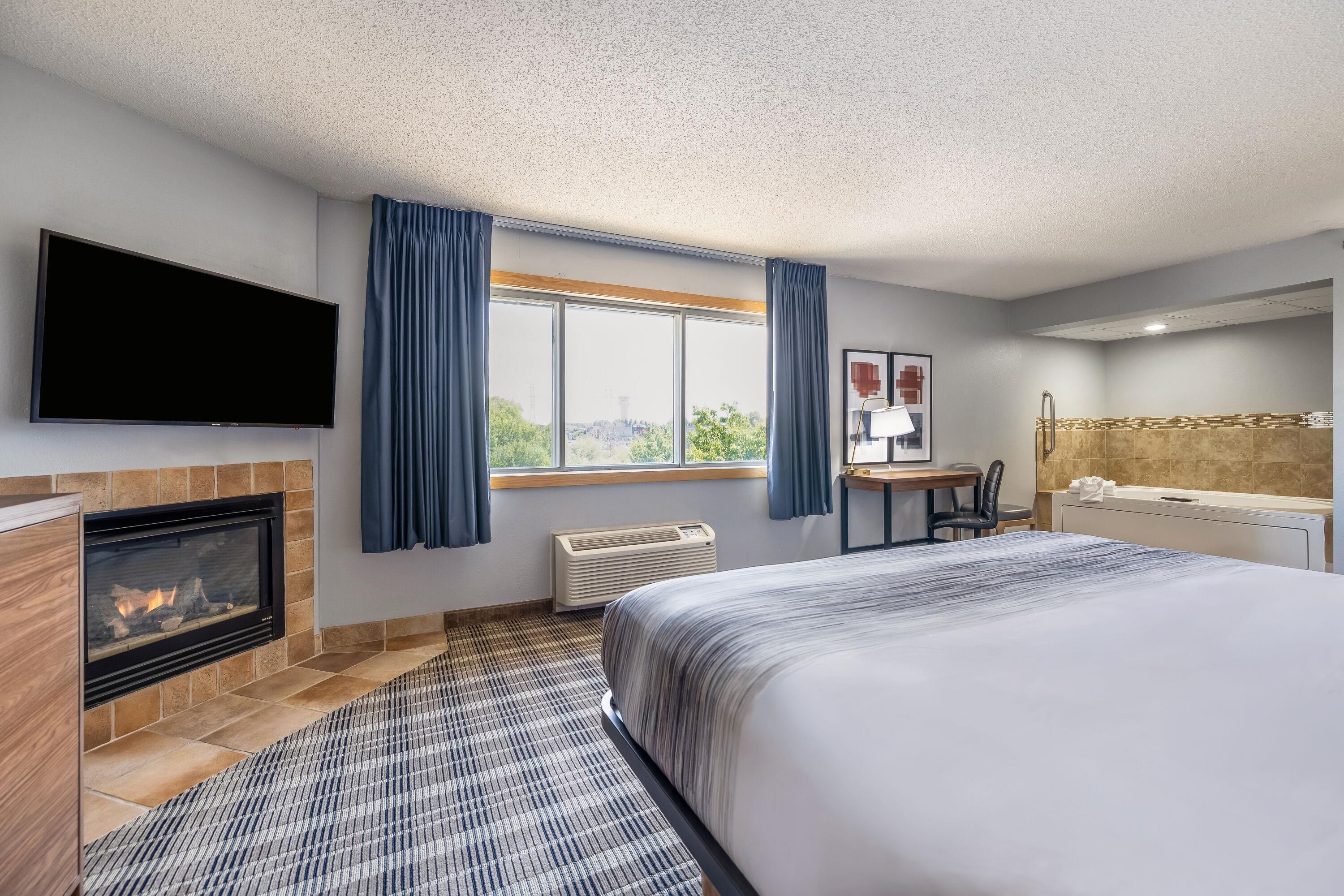Ein Hotelzimmer mit Kamin, einem Bett, einem Schreibtisch, einem Sessel, einem Fernseher, einem Fenster mit Vorhängen und einem Badezimmer.