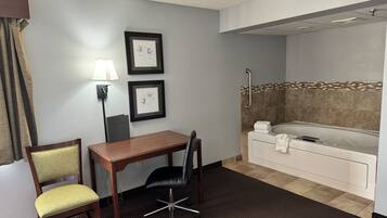 A hotel room with a desk, two chairs, a framed picture, a lamp, and a bathtub.