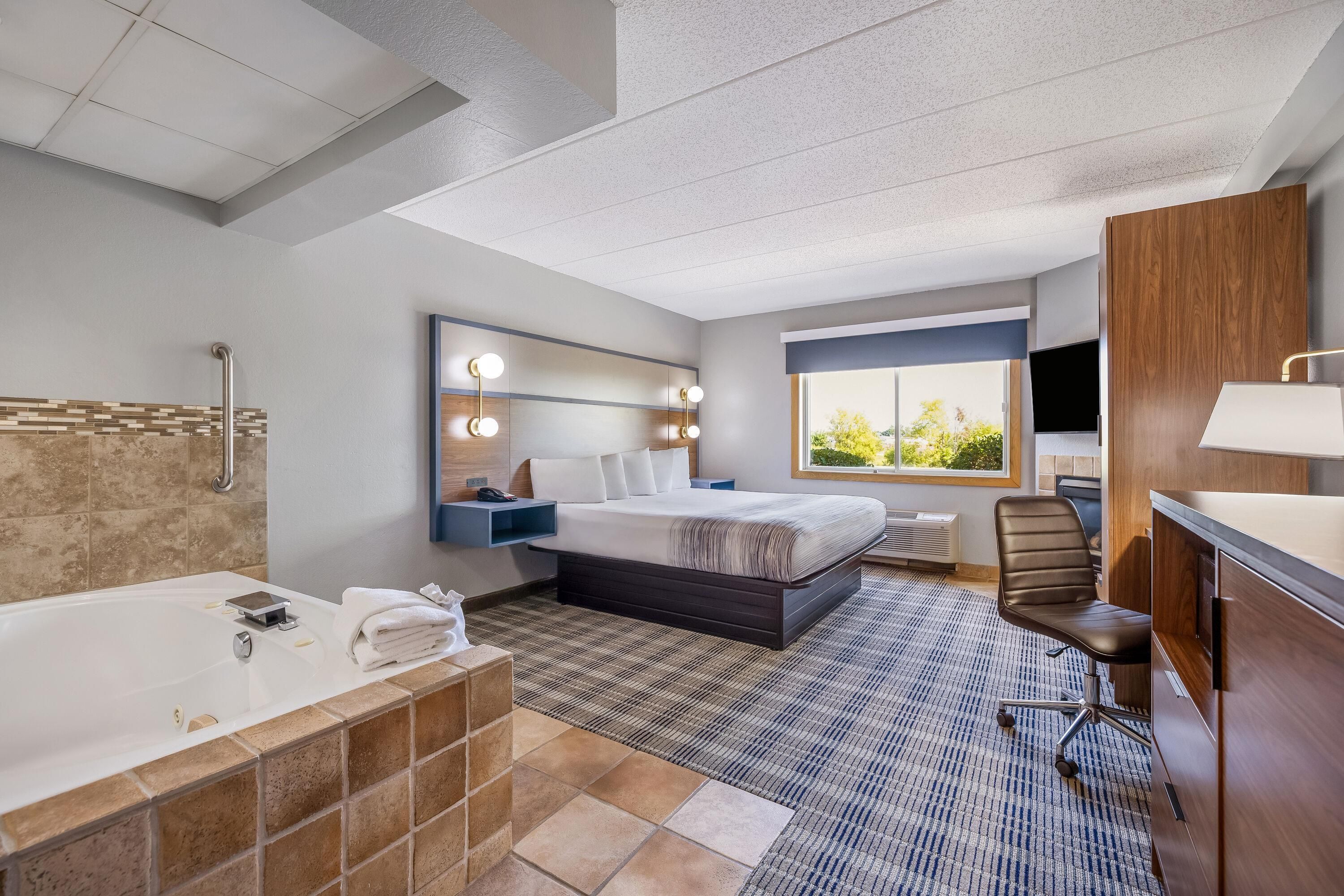 A hotel room with a large bed, a separate bathroom with a bathtub, a seating area with a chair, and a window with a view of greenery.