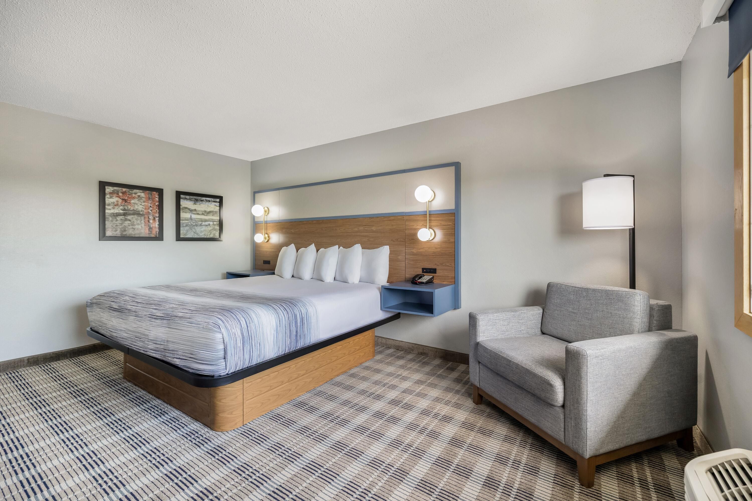 A hotel room with a bed, a chair, a bedside table, a lamp, and two framed pictures on the wall.