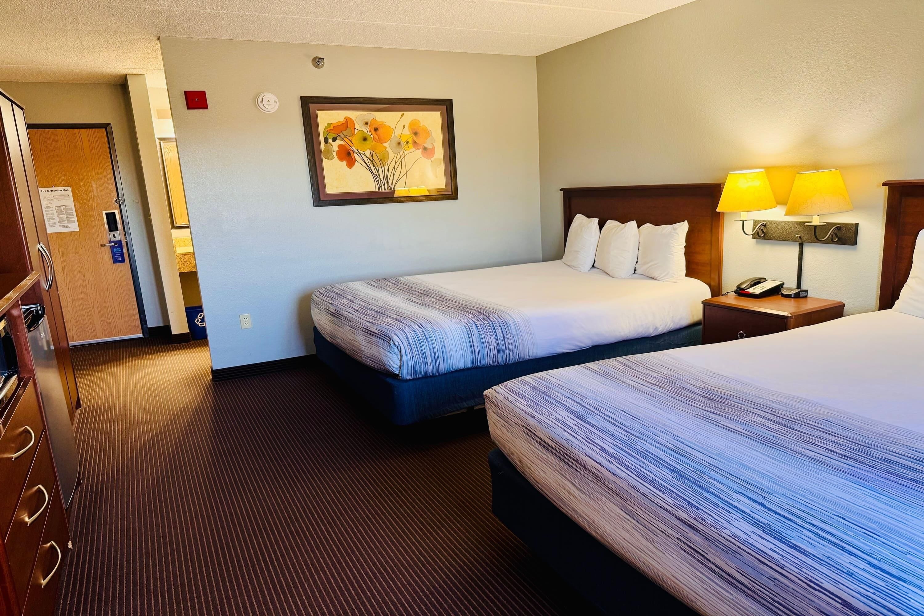 Quarto de hotel com duas camas, uma escrivaninha com luminária, um quadro na parede e uma porta com placa.