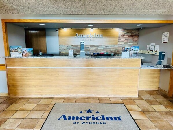 Un mostrador de recepción con el logotipo de AmericInn, una encimera de madera y una zona de recepción con suelo de baldosas.