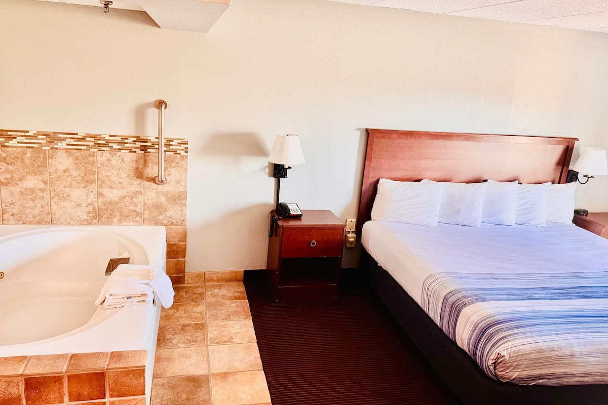 Hotelkamer met een groot bed, een aparte badkamer met badkuip, een nachtkastje van hout met een lamp en een telefoon aan de muur.