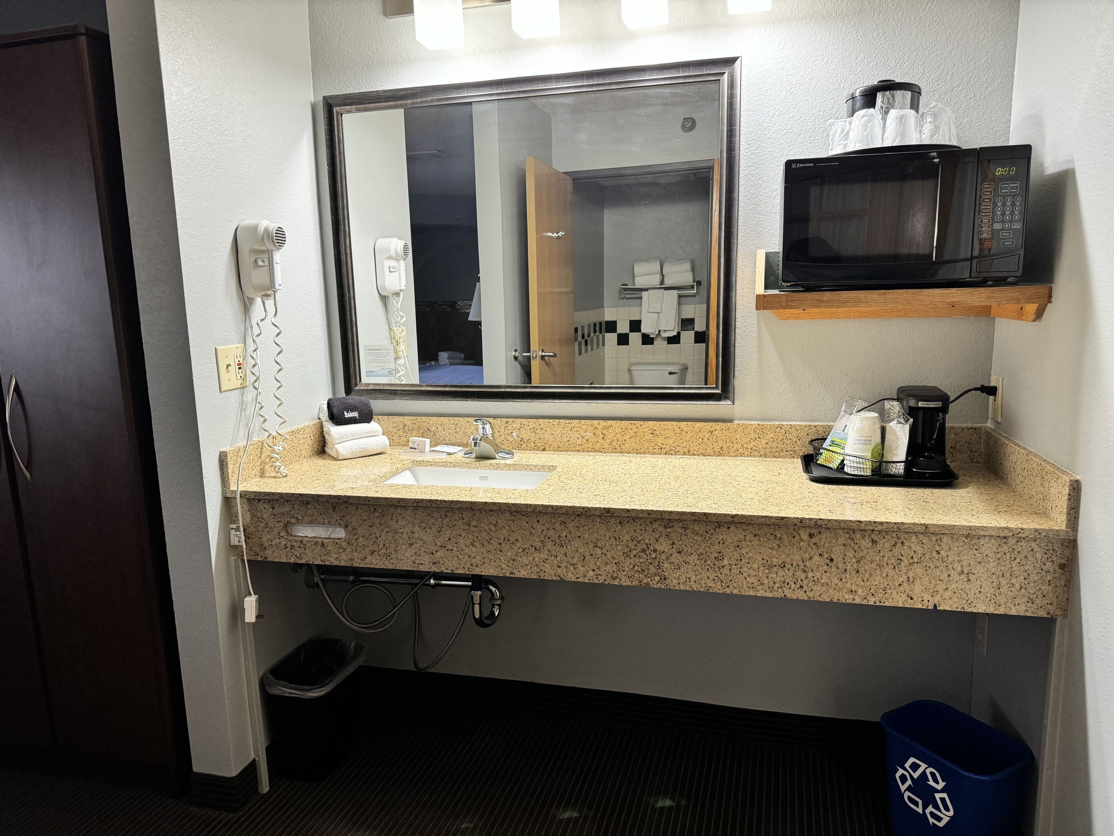 A hotel bathroom with a granite countertop sink, a mirror, a microwave, and a coffee maker.