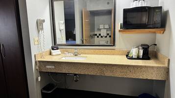 A hotel bathroom with a granite countertop sink, a mirror, a microwave, and a coffee maker.